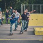 Een groep skateboarders racet over een verhard pad in een skatepark, omringd door toeschouwers. Ze naderen gele schansen en obstakels, terwijl een menigte toekijkt vanachter barrières en grasvelden. Op de achtergrond zijn bomen zichtbaar.