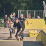 Drie mannen rijden op skateboards in de richting van de camera in een skatepark. Toeschouwers kijken toe op de achtergrond bij een gele schans en leuningen, met bomen en gras eromheen. De sfeer is levendig en ongedwongen.