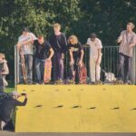 Vijf skateboarders staan bovenaan een gele schans, klaar om in te vallen, terwijl een fotograaf onderaan knielt om een foto van hen te maken. Verschillende mensen en kinderen kijken toe op de achtergrond in een zonnig park.