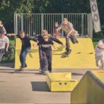 Een groep volwassenen, sommige hardlopend en sommige skateboardend, racet over een schans van een skatepark, met toeschouwers op de achtergrond op een zonnige dag.
