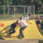 Drie skateboarders rijden samen op een gele schans met rode vlekken, terwijl verschillende mensen in de buurt toekijken en foto's nemen. Bomen en geparkeerde fietsen zijn zichtbaar op de achtergrond op een zonnige dag.