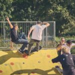 Twee skateboarders voeren trucs uit op een gele schans met rode vlekken, terwijl een groep mensen toekijkt en hen fotografeert in een buitenpark omringd door bomen.