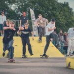 Een groep skateboarders racet over een gele schans in een openlucht skatepark, met toeschouwers op de achtergrond op een zonnige dag. Bomen en een menigte creëren een levendige sfeer.
