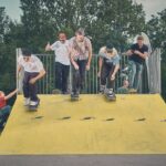 Vijf skateboarders racen van een gele schans in een park, terwijl toeschouwers, waaronder kinderen en volwassenen, vanaf de zijkant toekijken. Bomen en een bewolkte lucht zijn op de achtergrond te zien.