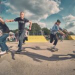 Vier skateboarders racen op een zonnige dag in een skatepark, met een menigte die toekijkt op de achtergrond en gele ramps zichtbaar onder een blauwe lucht met verspreide wolken.