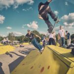 Een groep skateboarders in een skatepark met felgele schansen. Een skateboarder gaat de lucht in terwijl een andere een schans oprijdt. Verschillende mensen kijken toe onder een blauwe lucht met verspreide wolken.