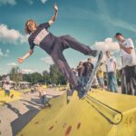 Een skateboarder met lang haar doet een truc op een gele schans in een skatepark, omringd door een groep mensen die toekijkt onder een blauwe lucht met verspreide wolken.