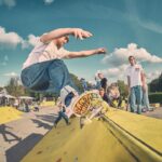 Een skateboarder in spijkerbroek en wit shirt doet een truc op een gele schans in een openlucht skatepark terwijl een groep mensen toekijkt onder een blauwe lucht met verspreide wolken.