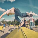 Een skateboarder valt midden in de lucht van een geel obstakel in een druk skatepark, terwijl anderen toekijken en skaten op de achtergrond onder een blauwe lucht met verspreide wolken.