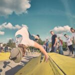 Een skateboarder voert een handplant-truc uit op een gele schans in een skatepark, omringd door toeschouwers en andere skaters onder een gedeeltelijk bewolkte hemel.