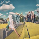 Een skateboarder doet een handstand truc op een gele skatehelling, terwijl een menigte mensen toekijkt onder een gedeeltelijk bewolkte hemel. Sommige toeschouwers staan dicht bij de schans, anderen staan op de achtergrond.