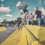 Een skateboarder doet een truc over een gele schans in een skatepark terwijl verschillende mensen met skateboards op de achtergrond toekijken onder een gedeeltelijk bewolkte hemel.