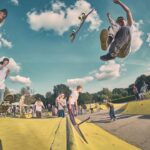 Een skateboarder in de lucht die een truc doet in een druk buiten-skatepark, terwijl anderen toekijken of vlakbij skaten onder een blauwe lucht met verspreide wolken.