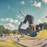 Een skateboarder voert een truc uit in de lucht in een openlucht skatepark, omringd door mensen die kijken en skaten. De lucht is gedeeltelijk bewolkt en op de achtergrond staan bomen.