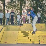 Een skateboarder doet een truc langs een gele leuning in een skatepark, terwijl een groep mensen met skateboards op de achtergrond toekijkt. Achter hen zijn bomen en groen te zien.