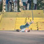 Een jonge skateboarder valt voorover op de stoep onderaan een gele trap in een skatepark, terwijl toeschouwers met skateboards toekijken vanaf de bovenkant van de trap.