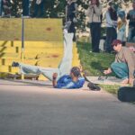 Een skateboarder in een blauw shirt valt midden in de lucht op de stoep bij gele schansen, terwijl toeschouwers en medeskaters op de achtergrond toekijken.