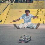 Een jongeman in lichtgekleurde kleding zit op de grond van een skatepark, met gestrekte benen, verbaasd te kijken. Zijn skateboard ligt voor hem op de stoep. Op de achtergrond zijn gele schansen en een zwart geschilderde rat te zien.