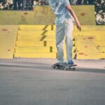 Een persoon in lichtgekleurde kleding rijdt op een skateboard bij gele schansen en trappen in een skatepark. Een ander skateboard ligt op de grond vlakbij. Bomen en mensen zijn op de achtergrond te zien.