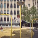 Een man met een hoed beklimt een gele straatlantaarn op een moderne binnenplaats met geometrische ramen, terwijl een andere persoon over de gele grond eronder loopt. Bomen en een groot gebouw staan op de achtergrond.