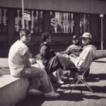 Vier jonge mannen zitten buiten te praten, twee op klapstoeltjes en twee op een laag betonnen muurtje, voor een modern gebouw met grote ramen. De scène is in zwart-wit.