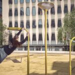 Een persoon schommelt met één arm aan een gele speeltuinpaal, benen gestrekt in de lucht, in een stedelijke buitenruimte met moderne gebouwen en bomen op de achtergrond.