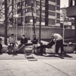 Een groep van zeven mensen zit en ontspant op een laag muurtje buiten moderne flatgebouwen; de een knipt de ander terwijl de anderen toekijken of wegkijken, waardoor een ongedwongen, stedelijk tafereel ontstaat.