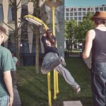 Een persoon springt om een gele straatlantaarn te pakken terwijl twee anderen toekijken. De scène is buiten in de buurt van moderne gebouwen, met bomen en gras op de achtergrond. De sfeer is ongedwongen en stedelijk.