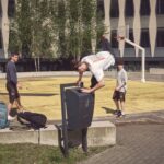 Een jongeman doet een handstand op een vuilnisbak bij een geel basketbalveld, terwijl twee anderen toekijken. Rugzakken en een waterfles liggen op de grond en op de achtergrond staat een gebouw met unieke ramen.