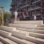 Een groep jongeren verzamelt zich rond moderne betonnen trappen in een stedelijk gebied. Eén persoon springt in de lucht van een richel terwijl anderen toekijken, met flatgebouwen en groen op de achtergrond.