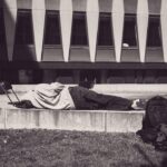 Een persoon ligt op een betonnen richel buiten een modern gebouw met geometrische ramen en gebruikt een rugzak en kleding als kussen. Twee lege stoelen en verspreide bezittingen liggen in de buurt. De scène is in zwart-wit.