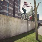Een persoon in de lucht springt van een hoge betonnen muur in de richting van een boom, terwijl een andere persoon op de muur in de achtergrond staat; moderne gebouwen en een wandelpad zijn zichtbaar in de buurt onder een heldere hemel.