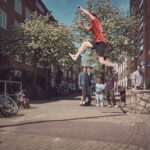 Een persoon in een rood shirt en een zwarte korte broek springt in de lucht over een straat in de stad, terwijl verschillende mensen toekijken en één een foto neemt op een zonnige dag. Langs de kant staan fietsen geparkeerd en op de achtergrond staan bomen.