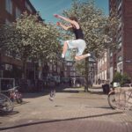 Een man in de lucht springt van een stenen richel in een straat in de stad terwijl een andere persoon in de buurt staat, een telefoon vasthoudt en hem lijkt aan te moedigen. Fietsen staan geparkeerd langs de straat; bomen bloeien op de achtergrond.