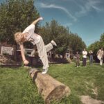Een persoon met krullend haar en vrijetijdskleding balanceert op een grote boomstam in een graspark, terwijl een groep mensen in de buurt toekijkt en foto's maakt. Bomen en gebouwen zijn zichtbaar op de achtergrond onder een blauwe lucht.