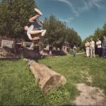 Een persoon maakt een backflip over een grote boomstam in een graspark, terwijl een groep mensen toekijkt onder een zonnige hemel en groene bomen.