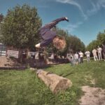 Een persoon met lang haar maakt een backflip over een boomstam in een graspark, terwijl een groep mensen in de buurt toekijkt. Op de achtergrond zijn bomen, gebouwen en een blauwe lucht te zien.