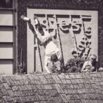 Een persoon voert een handstand uit op een stenen richel terwijl een ander in de buurt zit toe te kijken. Op de achtergrond zie je een muur met abstract reliëf en het woord "PopFest". De afbeelding is zwart-wit.