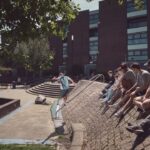 Een groep mensen zit op een schuine bakstenen muur en kijkt naar een persoon in lichtblauwe sportkleding die balanceert op een reling. Moderne gebouwen en bomen zijn zichtbaar op de achtergrond op een zonnige dag.