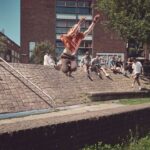 Een persoon in een oranje shirt springt door de lucht over een stenen richel in een stedelijk gebied terwijl een groep mensen toekijkt vanaf een grashelling op de achtergrond.