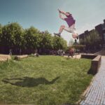 Een persoon in korte broek en een paars shirt maakt een atletische sprong in de buitenlucht, in de lucht boven een grasveld, met bomen en bakstenen gebouwen op de achtergrond onder een heldere hemel.