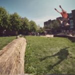Een persoon in de lucht springt over een grote houten boomstam in een graspark, met verschillende mensen en bakstenen gebouwen op de achtergrond onder een heldere hemel.