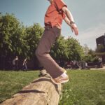Een persoon met lang haar loopt langs een grote omgevallen boomstam in een graspark op een zonnige dag, balancerend met uitgestrekte armen. Op de achtergrond zijn bomen en mensen te zien.