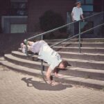 Een persoon in een wit shirt en korte broek balanceert ondersteboven op een groene leuning over betonnen treden, met één hand en beide voeten op de leuning. Andere mensen staan vlakbij en kijken toe.
