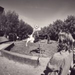 Een persoon maakt een salto boven een wegwijzer, gadegeslagen door een groep jongeren. Bomen en gebouwen omringen het grasveld en het beeld is zwart-wit.