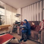 Een vrouw in een blauwe en grijze hijab zit op een beige bank een boek te lezen in een gezellige woonkamer, omringd door kussens, planken met spullen, een salontafel en een raam dat daglicht binnenlaat.