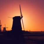 Silhouetten van drie traditionele windmolens staan tegen een levendige oranje en paarse zonsondergang, met bomen en een weg zichtbaar op de achtergrond.
