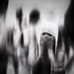 Een persoon staat in een wazige menigte en bedekt zijn gezicht met zijn handen. Het zwart-wit beeld heeft een bewegingsonscherpeffect en creëert een gevoel van verwarring of emotionele onrust.