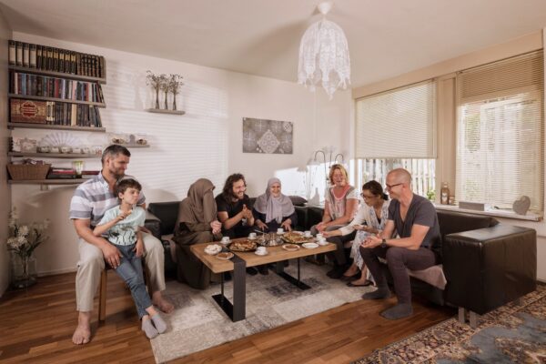 Een groep van zeven mensen, waaronder volwassenen en een kind, zitten samen in een gezellige woonkamer, delen hapjes en drankjes rond een koffietafel, glimlachen en genieten van elkaars gezelschap.