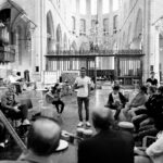 Een groep mensen zit in een grote, lichte kerk en vormt een cirkel. Sommigen spelen op muziekinstrumenten, terwijl een man in het midden staat te zingen of te spreken. De sfeer is levendig en samenwerkend.